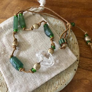 May include: A necklace featuring a tan leather cord strung with cylindrical green glass beads, round metallic beads, and a central clear faceted glass bead. The cord includes adjustable knots and small green accent beads throughout the design.