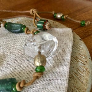 May include: A bracelet featuring a mix of clear glass, dark green glass, and metallic beads strung on a tan leather cord. The design includes a large, irregular clear glass bead, cylindrical dark green beads, and small green spacers with knots.