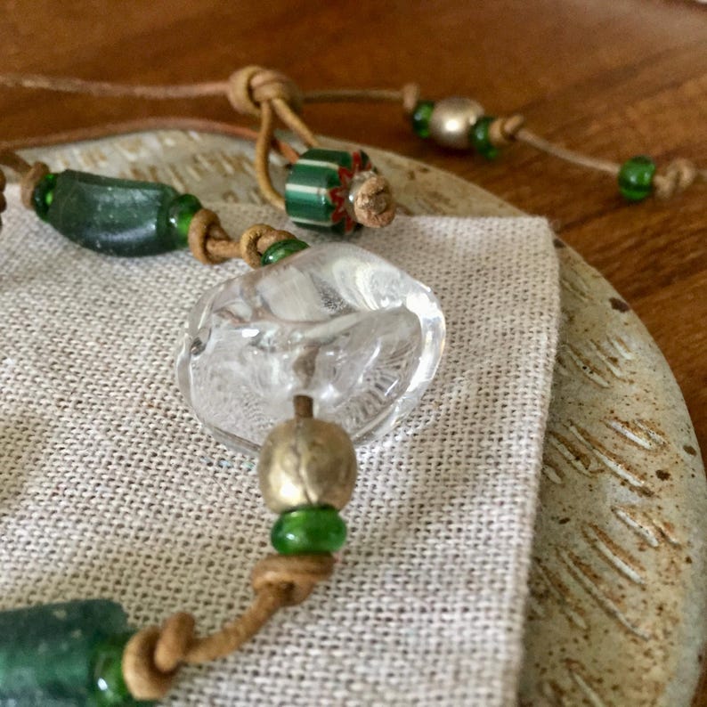 May include: A bracelet featuring a mix of clear glass, dark green glass, and metallic beads strung on a tan leather cord. The design includes a large, irregular clear glass bead, cylindrical dark green beads, and small green spacers with knots.