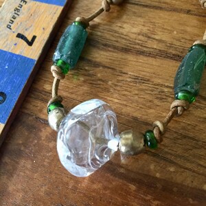 May include: A necklace featuring a tan leather cord strung with cylindrical teal glass beads, small green glass spacer beads, metallic beads, and a large clear textured glass bead, positioned next to a ruler showing 7 inches.