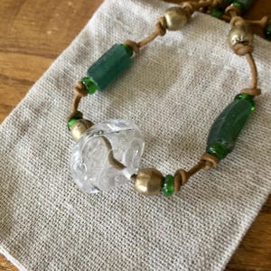 May include: A necklace featuring a tan leather cord strung with a clear, textured glass bead, cylindrical dark green glass beads, and round metallic beads. The beads are separated by small, vibrant green glass spacer beads along the cord.
