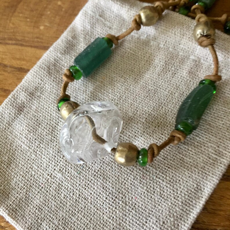 May include: A necklace featuring a tan leather cord strung with a clear, textured glass bead, cylindrical dark green glass beads, and round metallic beads. The beads are separated by small, vibrant green glass spacer beads along the cord.