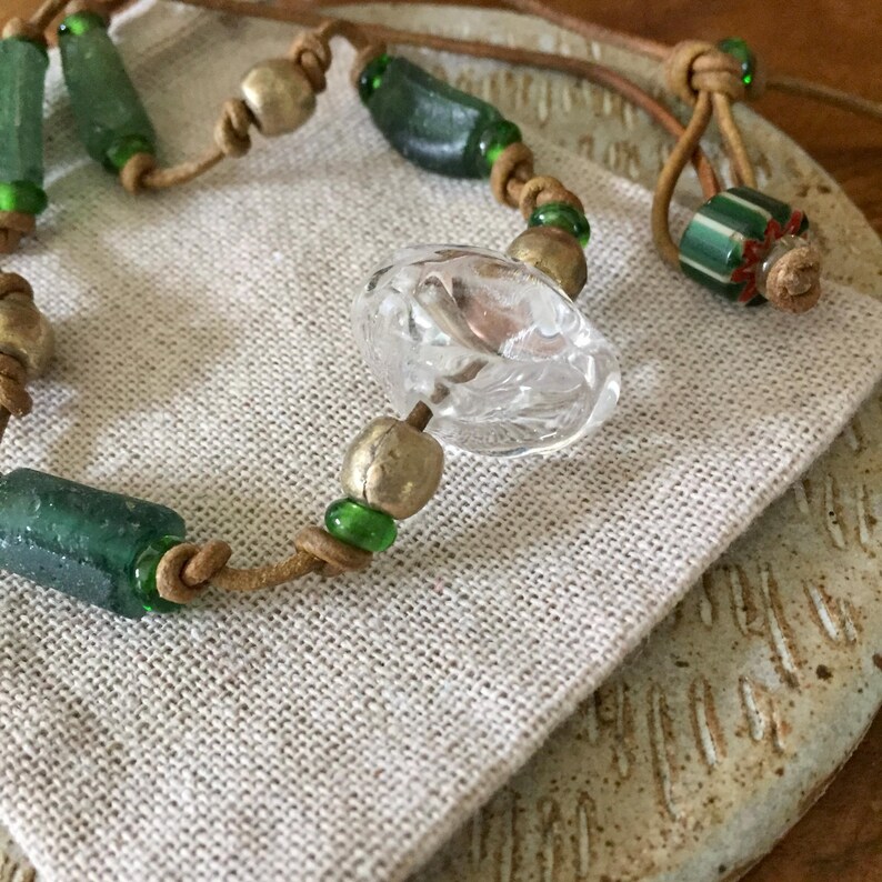 May include: A necklace featuring a tan leather cord strung with cylindrical dark green glass beads, round metallic beads, a clear faceted glass center bead, and a small green and white striped bead with red accents, all knotted in place.