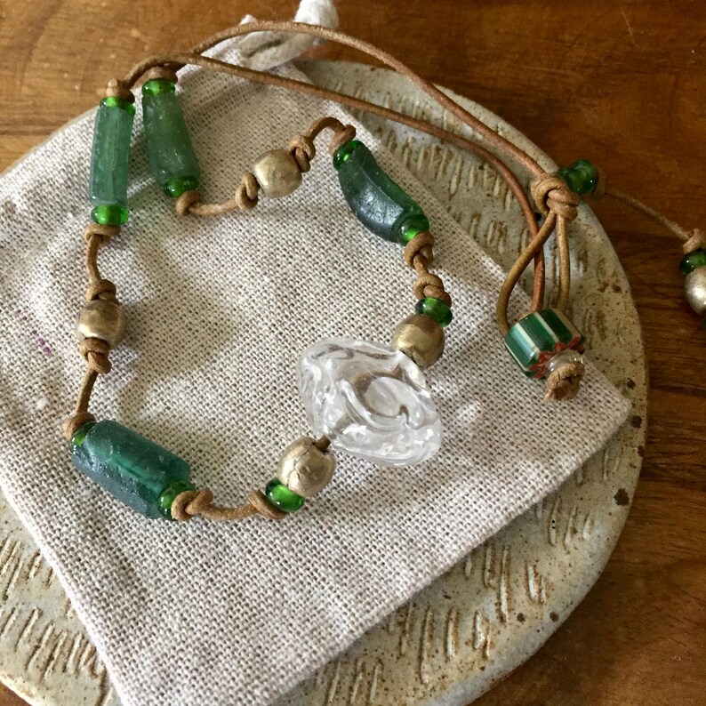 May include: A necklace featuring a tan leather cord strung with cylindrical green glass beads, round metallic beads, and a central clear, textured glass bead. The cord is knotted between the beads, and a striped green and white bead is near the end.