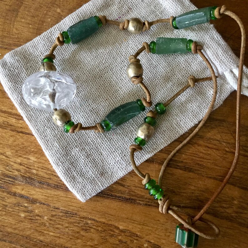 May include: A necklace featuring a tan leather cord strung with cylindrical green glass beads, small round metallic beads, and a single large, clear, textured glass bead. The beads are separated by knots along the cord, creating a structured design.