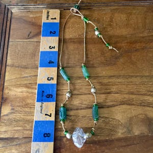May include: A necklace on a tan leather cord featuring green cylindrical beads, metallic spherical beads, and a central clear faceted bead. A wooden ruler is placed alongside the necklace, showing a total length of approximately 8 inches.