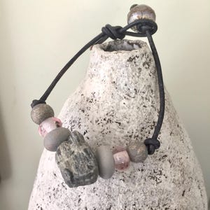 May include: A handmade bracelet featuring a mix of natural stone and glass beads in shades of gray, pink, and beige. The beads are strung on a black leather cord, with a knot closure. The bracelet is displayed on a textured, off-white surface.