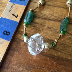 May include: A necklace featuring a central clear glass bead, flanked by metallic silver-toned beads and smaller green glass beads, strung on a tan cord. A wooden ruler is visible, showing the beads measure between 7 and 8 inches in length.