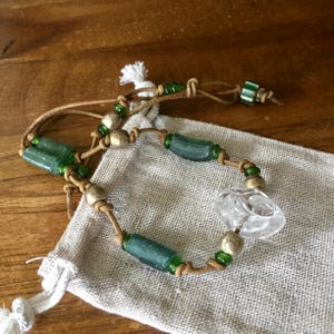 May include: A bracelet made of tan leather cord featuring cylindrical sea-glass beads in muted green, round metallic beads, small translucent green glass seed beads, and a single clear, faceted glass bead centerpiece with a knotted closure.