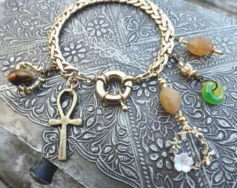May include: A gold-toned chain bracelet featuring a circular clasp and multiple charms, including a brass ankh, a small green ring, a white flower charm, and several smooth, rounded, earth-toned beads in shades of brown, tan, and amber.