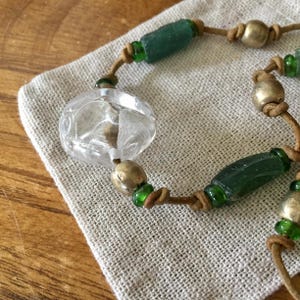 May include: A beaded necklace featuring a large clear glass bead with a textured surface, interspersed with cylindrical dark green glass beads, round metallic beads, and small bright green glass spacers strung on a tan leather cord.