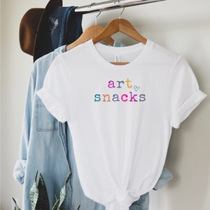 Puede incluir: Camiseta blanca con el texto "art ♥ snacks" en letras de colores.