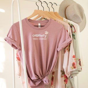May include: A dusty rose t-shirt with the text "creativity takes courage" and a small flower graphic.