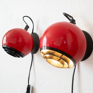 Puede incluir: Dos apliques de pared esféricos rojos con detalles negros e interior dorado. Cada lámpara tiene un cable y un enchufe negros. Las lámparas están montadas en una pared blanca.