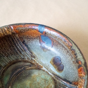 May include: Close-up of a handmade ceramic bowl with a textured rim and interior. The bowl features a blend of colors, including shades of green, brown, orange, and blue. The design includes textured patterns and a glossy finish.