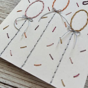May include: Hand-stitched birthday card featuring three embroidered balloons in pink, orange, and gold. The card has the words "Happy Birthday" stitched in purple thread, with confetti-like details.