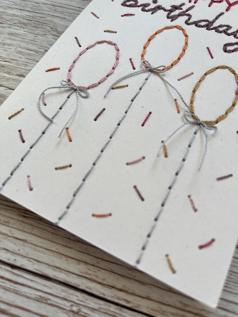 May include: Hand-stitched birthday card featuring three embroidered balloons in pink, orange, and gold. The card has the words "Happy Birthday" stitched in purple thread, with confetti-like details.