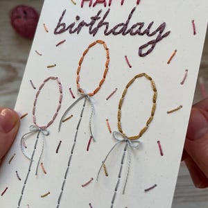 May include: A handmade birthday card with embroidered text and balloon designs. The words "HAPPY birthday" are stitched in red and purple thread. Three oval balloons are stitched in orange, pink, and gold, with gray strings and bows. Colorful confetti-like stitches decorate the white card.