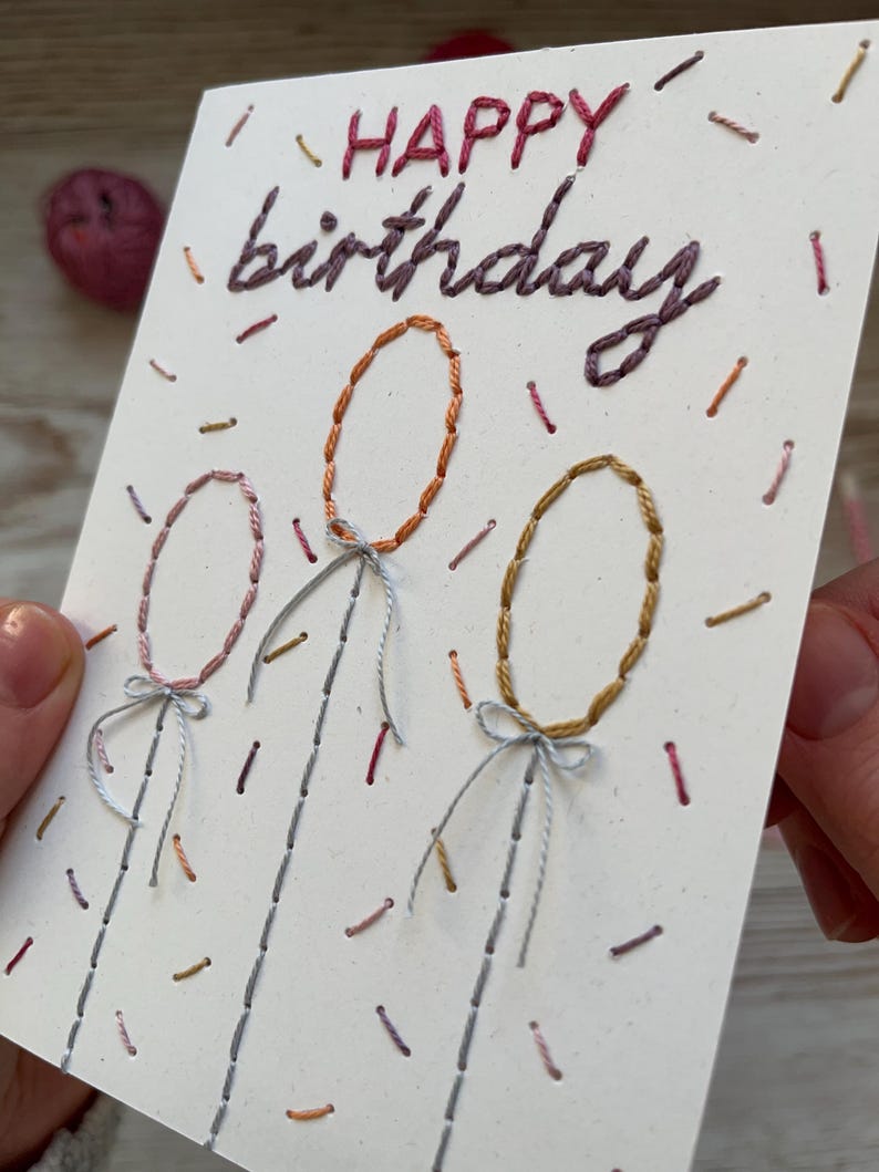 May include: A handmade birthday card with embroidered text and balloon designs. The words "HAPPY birthday" are stitched in red and purple thread. Three oval balloons are stitched in orange, pink, and gold, with gray strings and bows. Colorful confetti-like stitches decorate the white card.