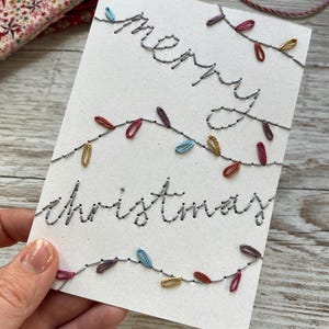 May include: A white greeting card featuring hand-stitched embroidery in grey thread that spells out merry christmas. The text is accented by embroidered string lights in various colours including red, blue, and gold, creating a festive design.