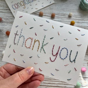 May include: A white rectangular greeting card featuring the hand-stitched text thank you in multicoloured embroidery thread. The card is decorated with scattered, short, straight embroidered stitches in various colours resembling sprinkles.