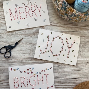 Op de afbeelding: Drie handgemaakte kerstkaarten met geborduurde tekst: "MERRY", "JOY" en "BRIGHT". De kaarten hebben rode en witte draad, met sneeuwvlok- en gloeilampaccenten. Een mand met garen en een schaar zijn ook zichtbaar.