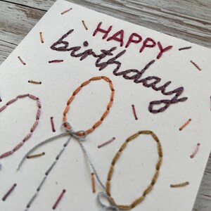 May include: Hand-stitched birthday card with the words "HAPPY birthday" in burgundy thread. The card features three embroidered balloons in shades of orange, pink, and gold, with gray string and bows. Small dashes of thread in various colors add to the festive design.