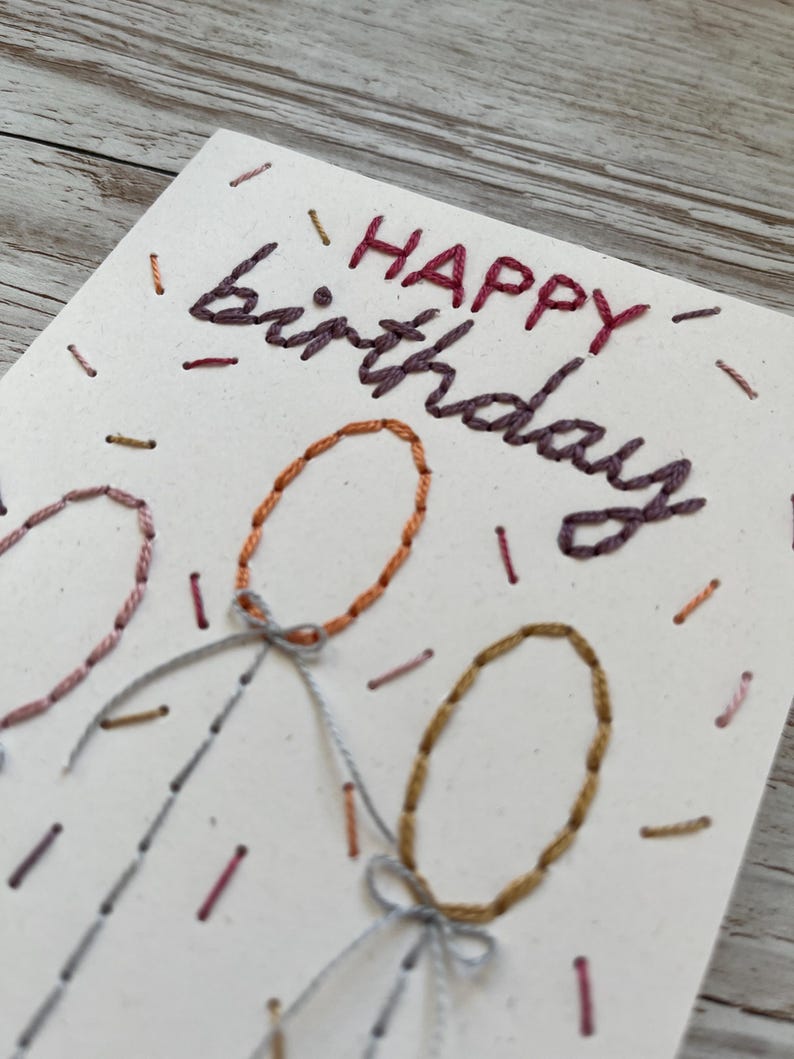 May include: Hand-stitched birthday card with the words "HAPPY birthday" in burgundy thread. The card features three embroidered balloons in shades of orange, pink, and gold, with gray string and bows. Small dashes of thread in various colors add to the festive design.