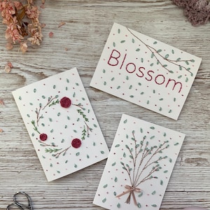 May include: Three white cards with embroidered floral designs and the word "Blossom". One card features a wreath of leaves and roses, another a branch with leaves, and the third a bouquet tied with a pink ribbon. Scattered pink and green dots.