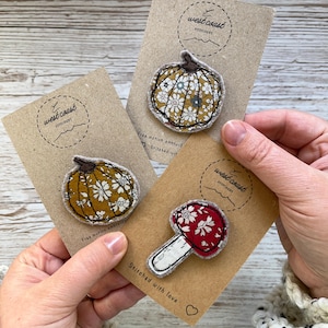 May include: Three fabric patches featuring two pumpkins and one mushroom, each attached to a brown card labelled west coast stitchery. The pumpkin patches have floral patterns in mustard and olive tones, while the mushroom has a red floral cap.