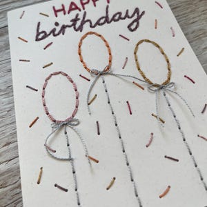 May include: Hand-stitched birthday card with the words "HAPPY birthday" in dark purple thread. The card features three embroidered balloon shapes in pink, orange, and gold, with gray string details. Small, colorful thread accents are scattered across the card.