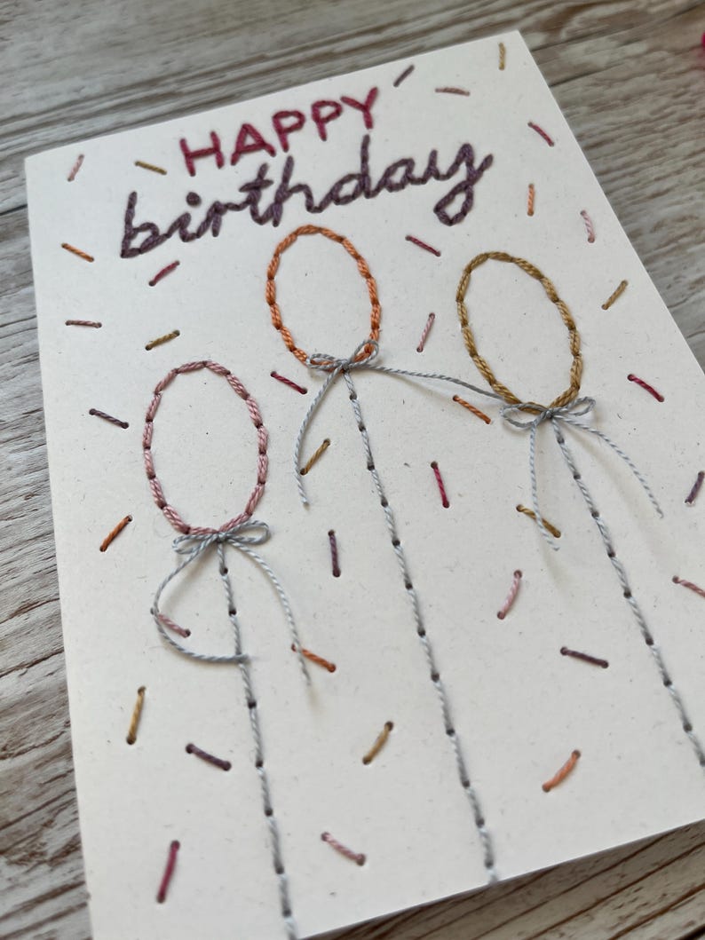 May include: Hand-stitched birthday card with the words "HAPPY birthday" in dark purple thread. The card features three embroidered balloon shapes in pink, orange, and gold, with gray string details. Small, colorful thread accents are scattered across the card.