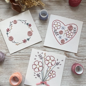 Peut inclure: Trois cartes carrées blanches ornées de broderies florales faites à la main dans des tons de rose, bleu et vert. Une carte présente une couronne circulaire, une autre un cœur rempli de fleurs et la troisième un bouquet noué d'un ruban rose.