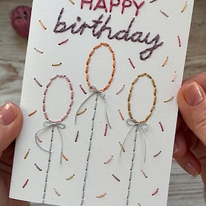 Può includere: Biglietto di compleanno fatto a mano con dettagli ricamati. La carta presenta le parole "HAPPY birthday" in filo viola e rosa. Tre forme di palloncini sono ricamate in arancione e oro, con cordini e fiocchi argentati. Punti colorati a forma di coriandoli sono sparsi sulla carta bianca.