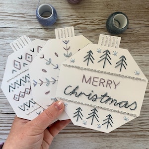 Pode incluir: Enfeites de Natal costurados à mão com desenhos bordados e a frase "Merry Christmas". Os enfeites são brancos com bordados prateados e azuis. Bobinas de linha e pequenas tesouras cor-de-rosa no fundo.