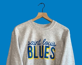 May include: A heather gray sweatshirt with the words "saint louis BLUES" printed on the front in blue and yellow. The sweatshirt is hanging on a wooden hanger against a blue background.