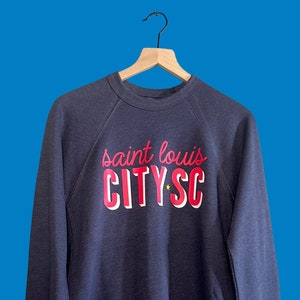 May include: A navy blue sweatshirt with the words "saint louis CITY SC" in a red and white design. The sweatshirt is hanging on a wooden hanger against a blue background. The text includes a star graphic.
