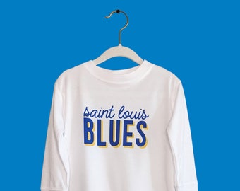May include: White long-sleeve shirt with the text "Saint Louis Blues" in blue and yellow. The shirt is hanging on a white hanger against a blue background. The text at the bottom says "comes in a bodysuit or a long sleeve shirt."