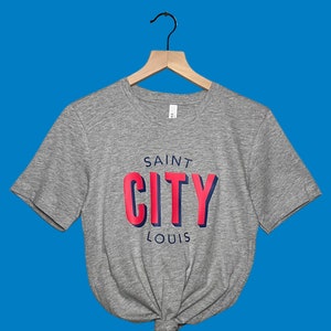 May include: A gray t-shirt with the words "SAINT CITY LOUIS" printed on the front in red and blue. The shirt is hanging on a wooden hanger against a blue background. The shirt is knotted at the waist.
