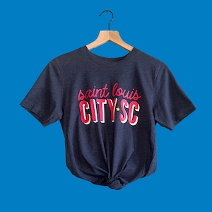 May include: A navy blue t-shirt with the text "saint louis CITY*SC" in red and white. The shirt is knotted at the waist and hangs on a wooden hanger against a blue background.