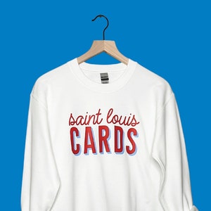 May include: White sweatshirt with the text "saint louis CARDS" in red script and block letters with a blue shadow. The sweatshirt is displayed on a wooden hanger against a blue background.