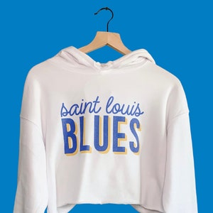 May include: White cropped hoodie with long sleeves, hanging on a wooden hanger against a blue background. The hoodie features the text "saint louis BLUES" in blue and yellow lettering, representing a sports team.