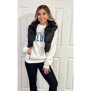 May include: A black puffer vest over a white sweatshirt with the text "Saint Louis BLUE" in blue and yellow. The person is wearing black leggings. The vest has a high collar and a zipper closure.