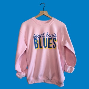 May include: A light pink sweatshirt with the text "saint louis BLUES" in blue and yellow lettering. The sweatshirt is hanging on a wooden hanger against a blue background. The sleeves are cuffed.