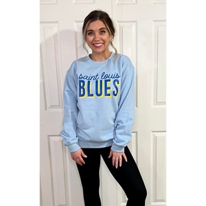 May include: Light blue sweatshirt with the text "Saint Louis BLUES" in blue and yellow. The sweatshirt is long-sleeved and has a crew neck. The person in the image is wearing black leggings.