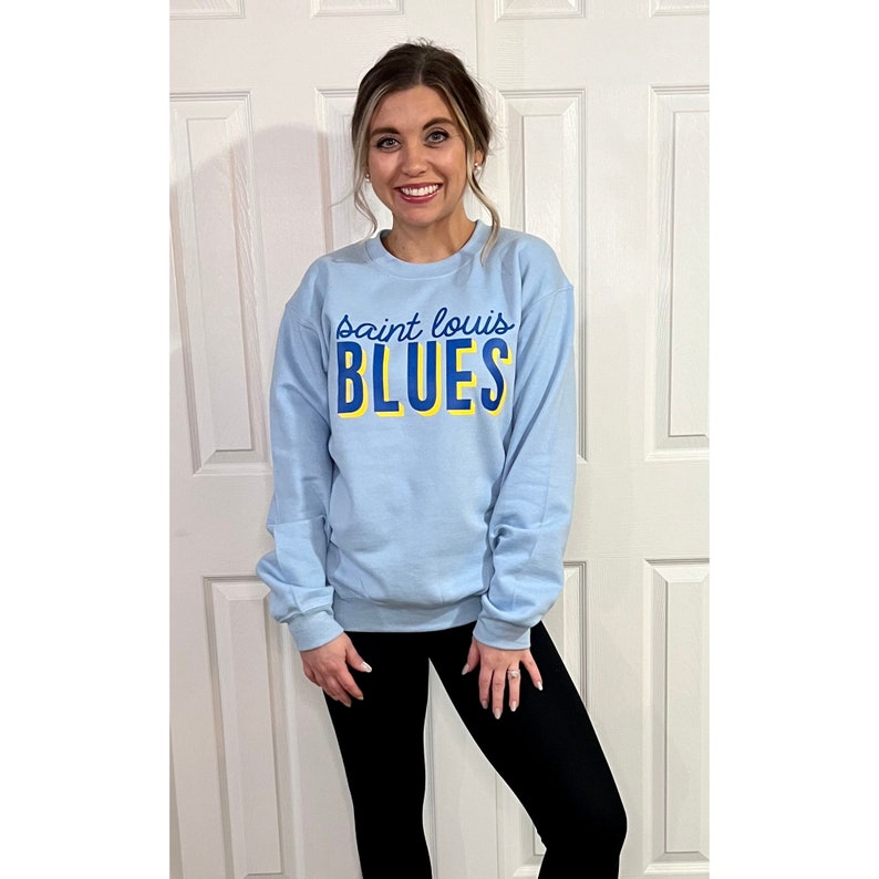 May include: Light blue sweatshirt with the text "Saint Louis BLUES" in blue and yellow. The sweatshirt is long-sleeved and has a crew neck. The person in the image is wearing black leggings.