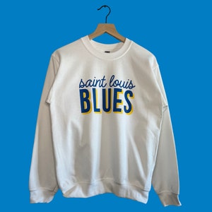 May include: White crewneck sweatshirt with the text "saint louis BLUES" in navy blue and yellow. The sweatshirt is hanging on a wooden hanger against a blue background. The sweatshirt is a casual style.