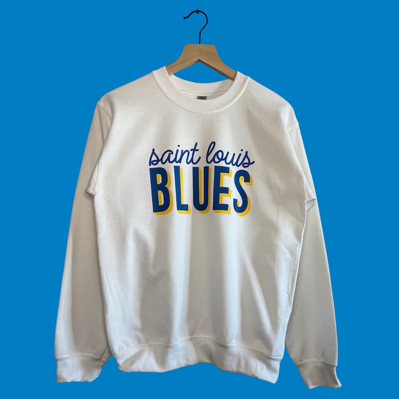 May include: White crewneck sweatshirt with the text "saint louis BLUES" in navy blue and yellow. The sweatshirt is hanging on a wooden hanger against a blue background. The sweatshirt is a casual style.