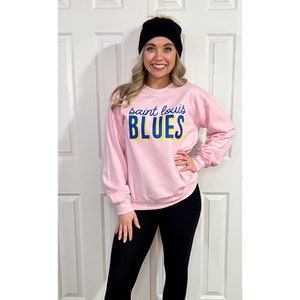 May include: A light pink sweatshirt with the text "saint louis BLUES" in blue and yellow. The sweatshirt is long-sleeved. The person in the image is wearing a black beanie and black leggings.
