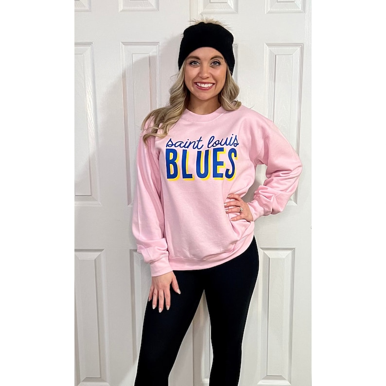 May include: A light pink sweatshirt with the text "saint louis BLUES" in blue and yellow. The sweatshirt is long-sleeved. The person in the image is wearing a black beanie and black leggings.